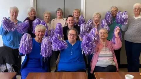 b:friend A group of older adults are gathered indoors in two rows, holding up purple pom‑poms. Several are seated at the front behind wooden tables, while others stand behind them. The group faces the camera in a bright room with neutral walls, and the purple pom‑poms create a coordinated, celebratory visual.