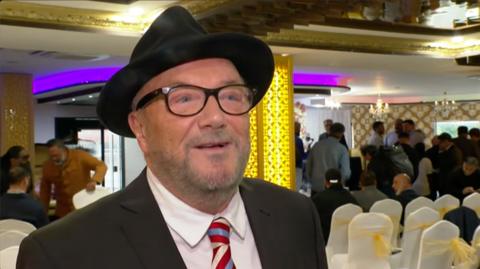 George Galloway wearing a black hat and a red, white and blue striped tie stands in front of a room full of white chairs with people talking to each other behind him.