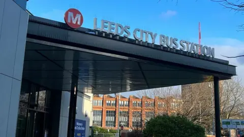 Hana Kelly/BBC Leeds city Bus Station sign on a triangular roof with black pillars down to the ground. In the background is a red brick building and above is a blue sky with fluffy clouds.