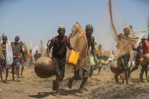 AFP via Getty Images Fishermen run towards the Matan Fada river. They're holding nets and gourds. One man in the front of the image can be seen holding a yellow jerrycan.