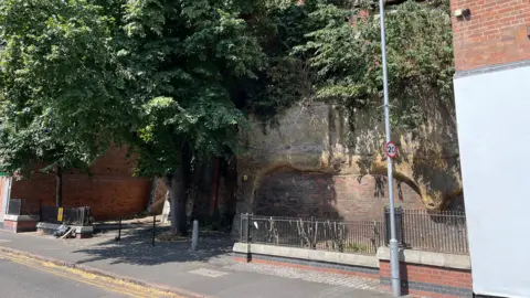 Area of Cliff Road where the cliff face has been cleared and meets the pavement area. Two buildings are visible either side.