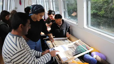 Ikon A woman standing up next to a young man who is sitting at a wooden table inside a narrowboat. The young man has black hair and glasses and is wearing a white and blue striped top. He is pointing at a loom-type piece of equipment, and there is colourful knitting wool behind it