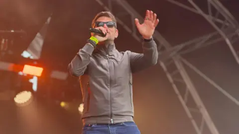 Roy Stride from Scouting for Girls - with a grey zipped jacket and dark sunglasses. He has the microphone in one hand and the other is raised.