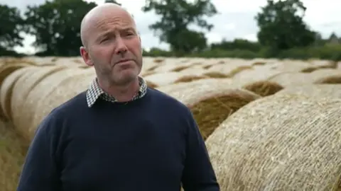 BBC A clean-shaven man in a blue jumper with a checked shirt underneath stood next to hay bales in a field