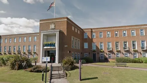 Royal Borough of Windsor and Maidenhead council building - a three-storey brick building, with a grand-looking entrance with a union flag flying over the top. There is a lawn in the foreground with some vegetation and benches in front