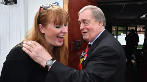 Angela Rayner and John Prescott laughing together on the campaign trail in 2017. Prescott has his hand on Rayner's shoulder. Rayner has glasses pushed back on her head.