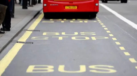A red bus is parked in a bus stop - yellow marking show a rectangular box on the road with the words "bus stop" repeated in it. The feet of passengers can be seen on a pavement to the left of the picture.