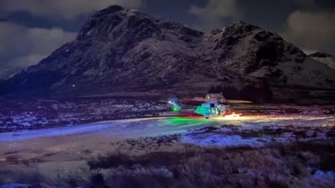Glencoe Mountain Rescue Team A helicopter sitting on snowy ground. Green and orange lights are coming from it. A large, dark hill is in the background and is partially covered with snow.
