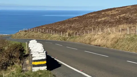 A section of the Mountain Road heading down towards Ramsey with the sea in the distance. there is a section of low stone wall painted white with yellow and black stripes in the side to the left and fencing on the hillside to the right of the road.