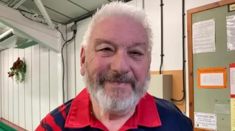 Mr Homer smiling at the camera, wearing a red and black polo shirt. He has white-grey hair and a grey beard and moustache, and is standing in a hall with an information board behind him and some sort of electrics box. The wall is panelled in white. 
