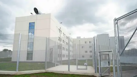 PA Media HMP Fosse Way is pictured behind a tall fence. It is a tall white-cladded building.