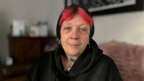 Martin Giles/BBC Sam Fairbairn wears a black hoodie and a black hat on her head. Her hair is red. She is looking sadly at the camera. She is sitting in a living room.