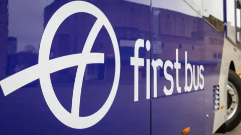 Part of a First Bus bus is pictured in close-up. It has the company name and logo in white on a blue background.