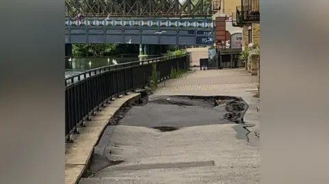 We Love Peterborough A tarmac pathway running beside a river which seems to be buckling and  has a small hole in the middle of it. A blue iron railway bridge can be seen in the background