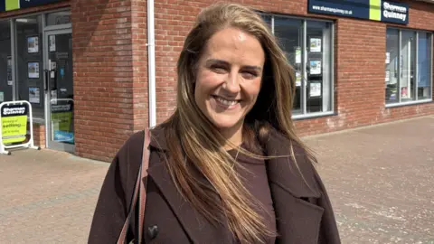 Ben Schofield/BBC Maria Banulus standing outside a shop, with long golden hair, wearing a brown top and coat, with a bag on one shoulder, and smiling at the camera. 