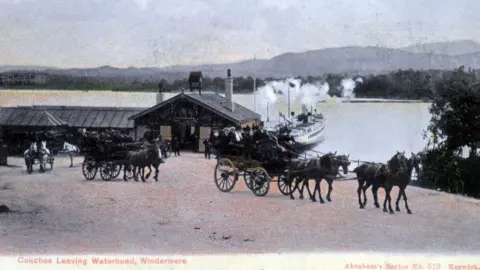 Windermere Lake Cruises Historic photo in black and white, tinged with sepia and pale pink on the ground, showing horse-drawn open coaches, filled with passengers. Behind them, a steamer is drawn up next to a ferry landing on the shore of Windermere. Mountains can be seen in the distance.