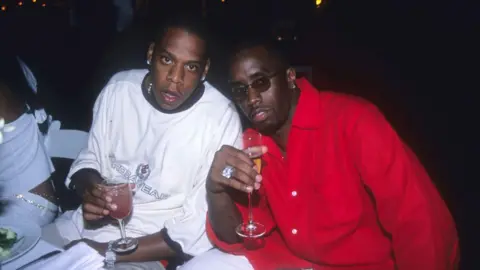 Getty Images Jay-Z e Sean "Diddy" Combs em um evento na casa do Sr. Combs em 2000. Jay-Z está sentado à esquerda em uma mesa segurando uma bebida. O Sr. Combs está sentado ao lado dele, também segurando uma bebida.