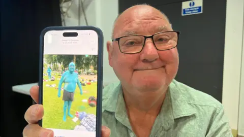 Close up shot of a bald man wearing glasses and a green shirt. He is holding a phone in front of him. On the phone is a picture of himself painted in blue wearing black shorts