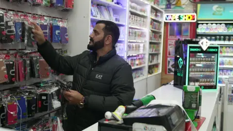 Amjad Khan is behind a shop counter and reaching up to hang up some phone cases that are on display behind him. In the background, various produce including vapes are on display. 