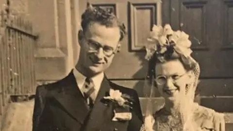 Family photo A black and white photo of a man with short, curly hair and glasses, wearing a black suit, white shirt and a tie. He has a flower pinned to his jacket and is smiling at the camera. Next t him is a woman wearing a white lacey dress, a white veil and a head piece and glasses and is also smiling at the camera.