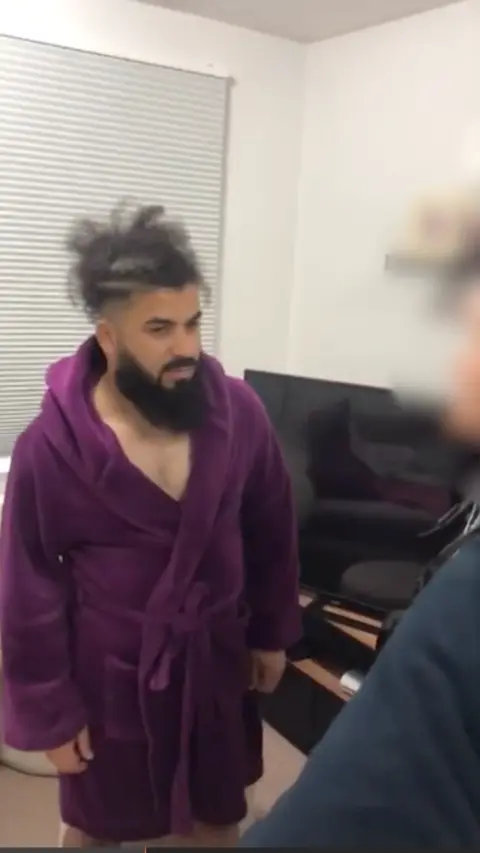NCA Man with fluffy black hair and a black beard wearing a purple dressing gown stood in front of a TV