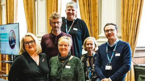 Norfolk and Suffolk NHS Foundation Trust A group of six people all looking towards the camera. There are four women and two men in the group. They are stood on a stage with some yellow curtains behind them. 