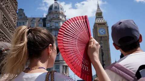 A close-up, over-the-shoulder shot from behind a woman with blonde hair in a ponytail holding up an open red hand fan. Next to her, a man wears a grey baseball cap. In the blurred background, London's Elizabeth clock tower rises against a clear blue sky on a bright, sunny day.