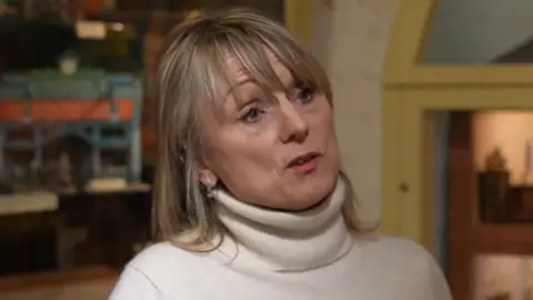 A woman with, long, blonde hair and a fringe. She is wearing a white turtleneck jumper.