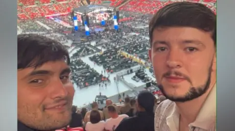 Connor Mann Ashwin and Connor take a selfie style image and smile. They are sat in the seats of a large arena. In the middle of it a boxing ring can be seen surrounded by seats.