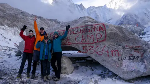 The family of four are standing with their arms in the air in-front of a big boulder which has red writing on that reads Everest Base Camp 5364m.