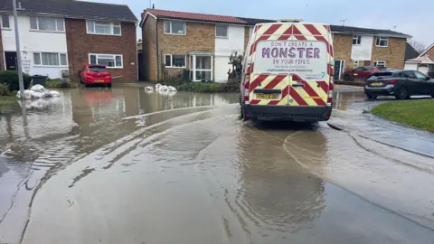 Stephen Huntley/BBC A water company van is parked on a flooded road. There are rubbish bags in the water in front of houses and cars parked in the background.