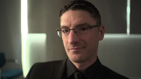 Mark Collett, who set up the far-right Patriotic Alternative, is pictured looking into the camera with a neutral expression, wearing glasses, a black shirt, black tie and black blazer.