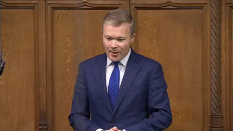 Bromsgrove MP Bradley Thomas stands in the House of Commons with the traditional wooden paneling behind him. He is wearing a navy suit with a blue tie and a white shirt.