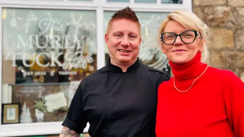 Photo of Lukasz and Edyta Kranz outside their cafe Murly Tuck in Tarves, he is wearing a black chef's top and she is wearing a red top.