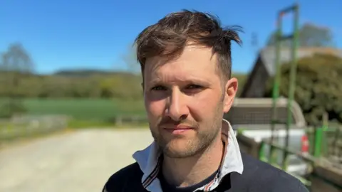 A man with short brown hair looks solemnly into the camera while wear a navy blue top with a white collar. Behind him is farm land and a trailer.