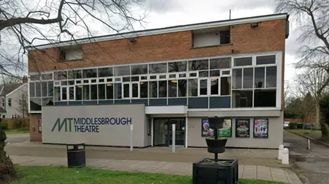 Google Middlesbrough Theatre is a three-storey standalone red-bricked building with a grey concrete front on the bottom floor. There are four show posters on display on the right of the exterior. Text reading "MT Middlesbrough Theatre" is painted on the left.