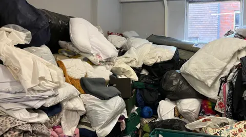 Tom Jackson/BBC Blankets, duvets and bed linen are all piled up in a small room which is painted grey and has a window on the right. 