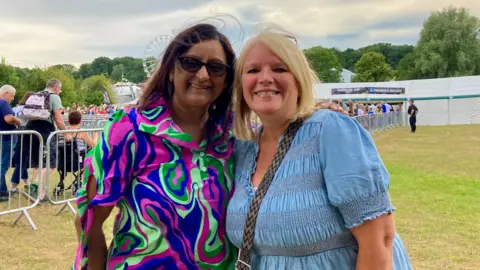 Two women standing in a field at a festival. They are both smiling and wearing dresses. The woman on the left has glasses on and a blue, green and purple patterned dress. She has her arm around the woman next to her, her friend, who is wearing a pale denim dress and has a cross-body bag over her chest.