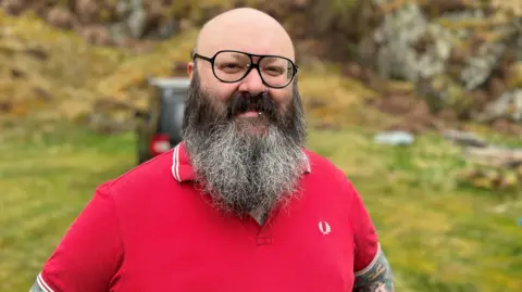 A bald-headed man with a dark beard and glasses, wearing a red top, looks at the camera with a rockface behind him