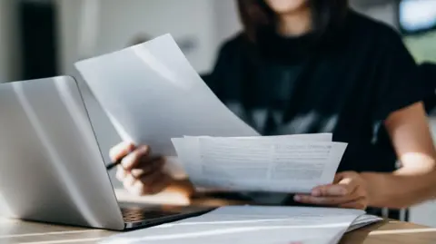 Getty A woman sits holding paperwork in front of a laptop.