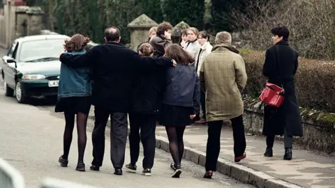 Getty Images A group of three teenage girls walk away from us down a stone-walled road. A man between them has his arms protectively draped over their shoulders. Others stand in groups beyond them. A man and a woman are also beside them, walking away.