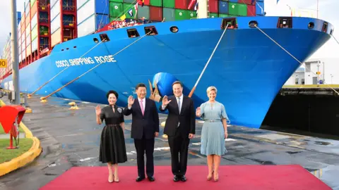 Getty Images Presiden Tiongkok Xi Jinping dan Presiden Panama Juan Carlos Varela, mengenakan jas dan dasi berwarna gelap, berdiri di antara Ibu Negara dengan pakaian formal, di depan kapal kontainer besar Tiongkok berwarna biru di pintu air Cocoli di Terusan Panama, pada 3 Desember 2018.