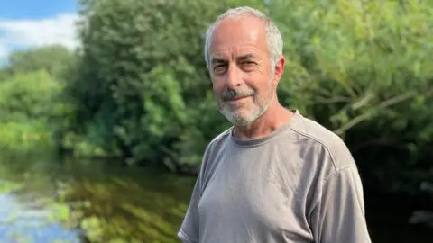Owen Sennitt/BBC A man wearing a grey shirt looks towards the camera, with a stretch of river visible in the background.