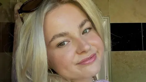 Saskia A young woman with blonde hair and a nose ring looking at the camera with bathroom tiles in the background.