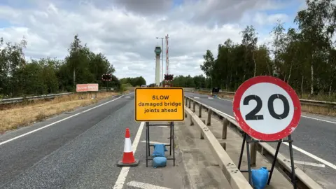 BBC/RICHARD EDWARDS This picture shows a yellow warning sign and a red and white speed limit sign warning of the 20mph restrictions over Selby bypass bridge. The green painted tower - which contains the controls to make the bridge swing open - is in the middle of the image.
