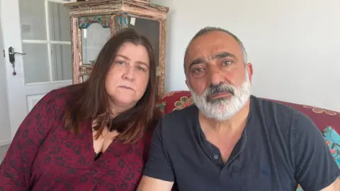 A woman with long brown hair wearing a purple v neck top sitting on a large armchair beside a man with a grey beard and grey hair wearing a grey t-shirt