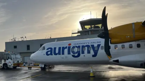 An Aurigny ATR aircraft on the tarmac, with the control tower in the background. The plane is white and yellow with propellers.