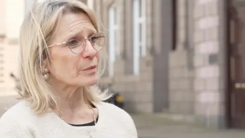 A woman with blonde hair and glasses is standing outside a building which is blurred in the background.