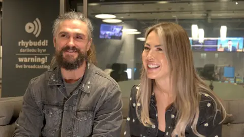 Lewis and Toni Prothero are smiling as they sit on a sofa in the BBC Wales building. Signs saying 'warning! live broadcasting' and 'rhybudd! darlledu byw' can be seen in the background.