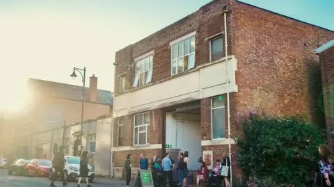 Two Queens Artists sitting and standing outside the gallery - a two storey brick building/warehouse